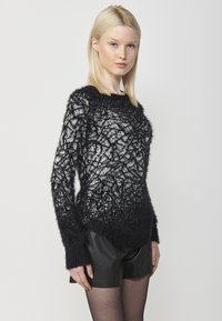 Pullover soffice nero e grigio con un motivo a foglia testurizzato, scollo rotondo, maniche lunghe e orlo posteriore leggermente più lungo, indossato sopra pantaloni corti neri.