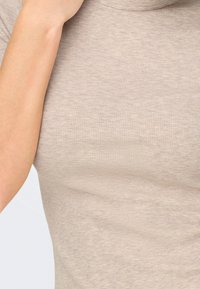 Gros plan d'une personne portant un haut en maille beige côtelé à manches courtes, avec un bras plié près du cou.