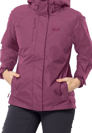 Donna che indossa una giacca impermeabile Jack Wolfskin viola con le mani nelle tasche laterali, abbinata a pantaloni neri.