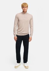 Ljus beige stickad tröja med zip-krage, matchad med mörka byxor och vita sneakers, vilket skapar en modern, avslappnad look.