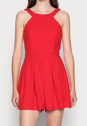 Ärmelloser roter Playsuit mit breiten Schulterträgern, tailliertem Oberteil und plissierten Shorts, getragen von einer stehenden Person vor neutralem Hintergrund.