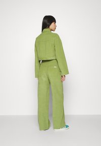 Geknipte groene corduroy jas met brede mouwen, gecombineerd met bijpassende hoge brede broeken. Heeft eenvoudige achterzakken en stiksels.