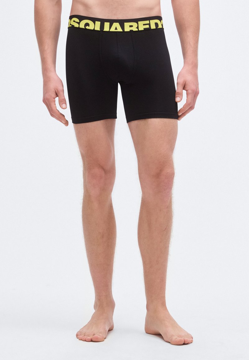 Bóxers negros hechos de tela elástica con una cintura ancha de logo amarillo. Presentan un diseño ajustado y una longitud hasta la mitad del muslo.