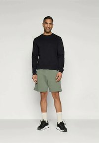 Μαύρο μακρυμάνικο πουκάμισο συνδυασμένο με ελιά κίτρινου σορτς. Το μοντέλο φοράει μαύρα αθλητικά παπούτσια και λευκές κάλτσες. Απλός σχεδιασμός, χαλαρή εφαρμογή.