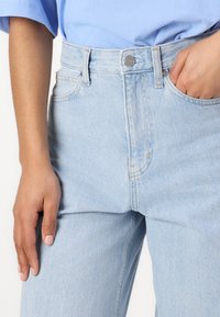 Ljusblå denimjeans med hög midja, som har knäppning med knappar, framfickor och en slät yta. Handen placerad på höften.