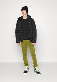 Black puffer jacket with a hood, worn over a black shirt, paired with olive green cargo pants and black high-top sneakers with mint accents.
