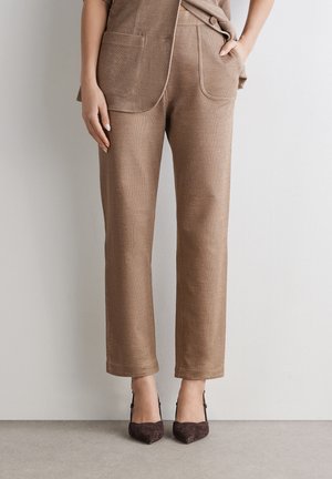 Frau trägt hellbraune, maßgeschneiderte Hosen mit großen Fronttaschen und dunkelbraune Slingback-Pumps mit spitzer Zehe, steht vor einer hellen Wand.
