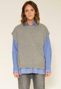 Gilet en tricot gris superposé sur une chemise à rayures bleues et blanches, avec un col arrondi et des manches courtes, associé à un jean foncé.