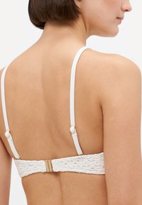 Biely čipkovaný podprsenka s textúrovaným vzorom, tenkými nastaviteľnými ramienkami a zlatými kovovými detailmi. Má zapínanie na háčik na zadnej strane.