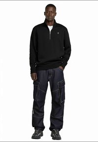 Black zip-up sweater, blue cargo pants with multiple pockets, and chunky black sneakers. The sweater has a stand-up collar and subtle branding.