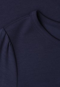 Gros plan sur un tissu bleu marine montrant un col rond et un détail froncé sur la couture d'épaule d'un vêtement.