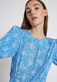 Bluza s cvjetnim uzorkom u plavoj boji, s napuhanim rukavima. Napravljena od laganog materijala; ima okrugli ovratnik i suptilnu prozirnu teksturu.