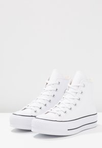 White high-top sneakers made of canvas with lace-up design, rubber sole, black stripe, and metal eyelets. Smooth texture, simple style.