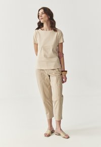 Beige top met korte mouwen en een vierkante halslijn, gecombineerd met beige cropped broeken. Het model draagt lichtgekleurde slide sandalen en amberkleurige armbanden.