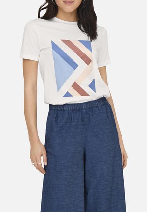 Femme portant un t-shirt blanc avec un imprimé géométrique bleu, beige et marron, associé à un pantalon ample bleu foncé avec une ceinture élastique.