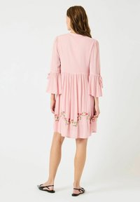 Abito rosa al ginocchio in tessuto testurizzato, con maniche a campana e ricami floreali sull'orlo. I lacci sulle maniche arricchiscono il dettaglio del design.