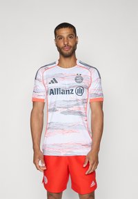 Jersey bianco a maniche corte con motivi astratti rosa e grigi, dettagli rossi e branding nero; abbinato a pantaloni corti rossi con un logo.