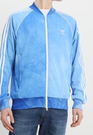 Mężczyzna ubrany w jasnoniebieską kurtkę zapinaną na zamek marki Adidas, z białymi paskami na rękawach i logo na piersi, w połączeniu z ciemnoniebieskimi dżinsami.