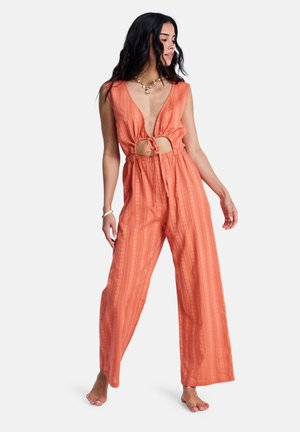 Žena stojící bosá, oblečená v oranžovém overal bez rukávů s hlubokým výstřihem do V a předním zavázáním, hledící stranou na bílém pozadí.