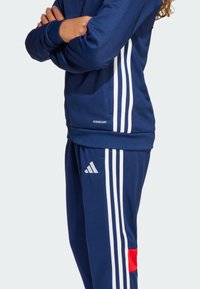 Chaqueta de chándal azul marino con rayas blancas y logo 'AEROREADY', combinada con pantalones a juego que presentan un logo de Adidas y un detalle en rojo.
