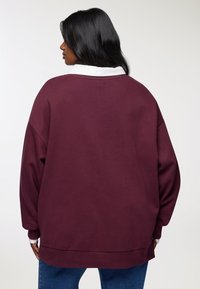 Kobieta z długimi czarnymi włosami, nosząca luźną bordową bluzę na białej koszuli z kołnierzykiem i niebieskich dżinsach, stojąca tyłem na jednolitym tle.