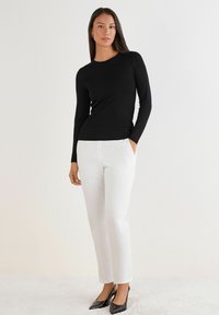 Schwarzes Langarmoberteil aus glattem Material, kombiniert mit weißen, maßgeschneiderten Hosen und schwarzen spitzen High Heels, was einen eleganten Look schafft.