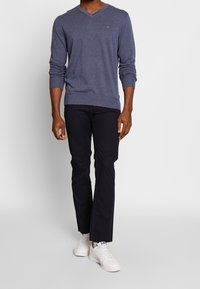 Pull col en V bleu marine, manches longues, texture lisse, associé à un pantalon noir et des baskets blanches. Détail à rayures sur la poitrine du pull.