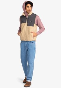 Beige Fleeceweste mit grauem Einsatz, mit Reißverschlusstaschen und hohem Kragen über einem rosa Kapuzenpullover; blaue Jeans; braune Stiefel.