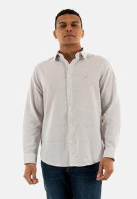 Camicia a righe bianche e navy a maniche lunghe con colletto button-down, logo sottile sul petto e texture del tessuto visibilmente morbida, abbinata a jeans scuri.