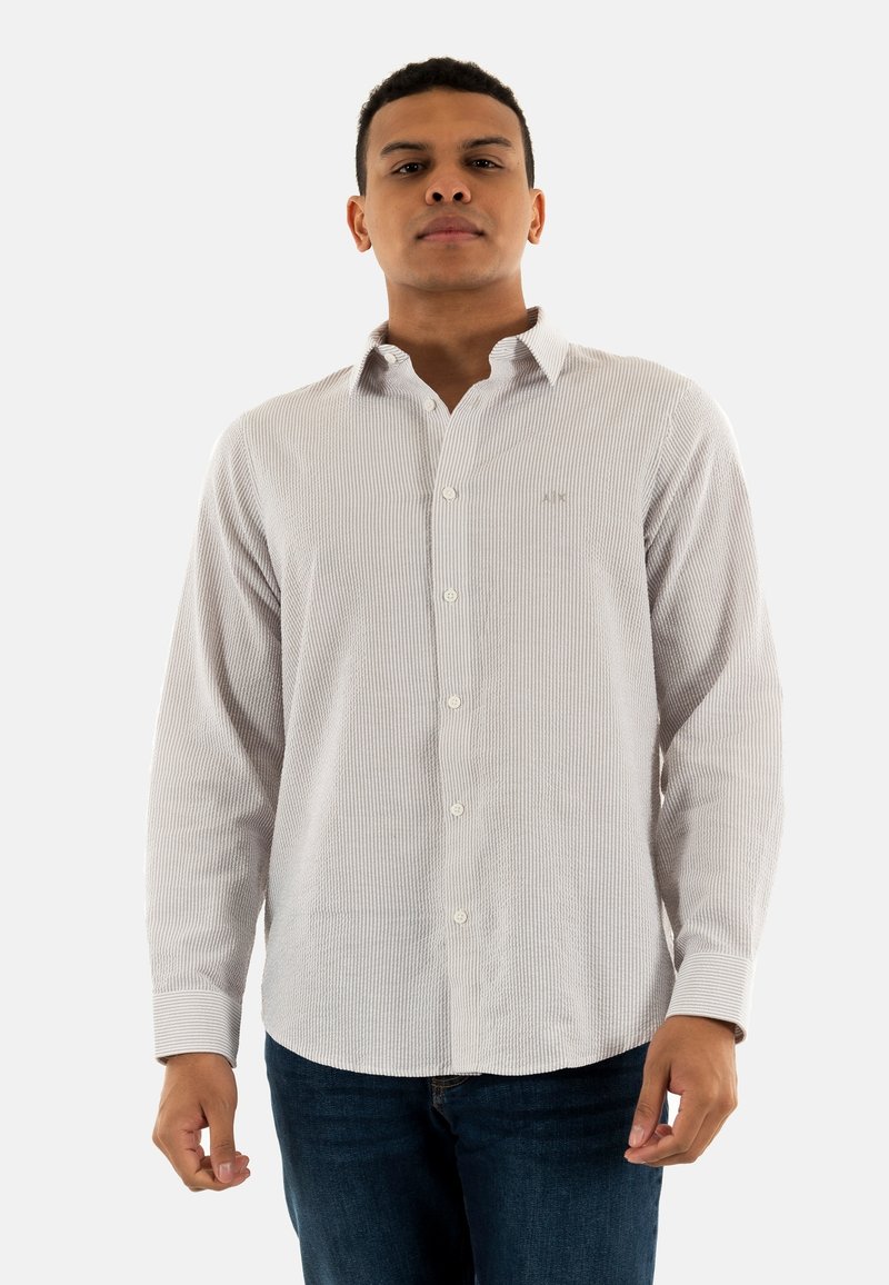 Camicia a righe bianche e navy a maniche lunghe con colletto button-down, logo sottile sul petto e texture del tessuto visibilmente morbida, abbinata a jeans scuri.