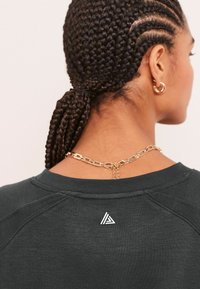 Tumehall sweatshirt ribatakaelusega, millel on seljaosas väike valge kolmnurgne logo. Juuksed on prantsuse põimitud, kaelakeel on kuldne ahel.