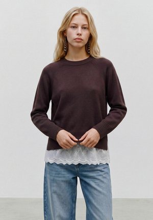 Jeune femme aux longs cheveux blonds porte un pull marron foncé par-dessus un top blanc en dentelle et un jean bleu, debout devant un fond clair uni.
