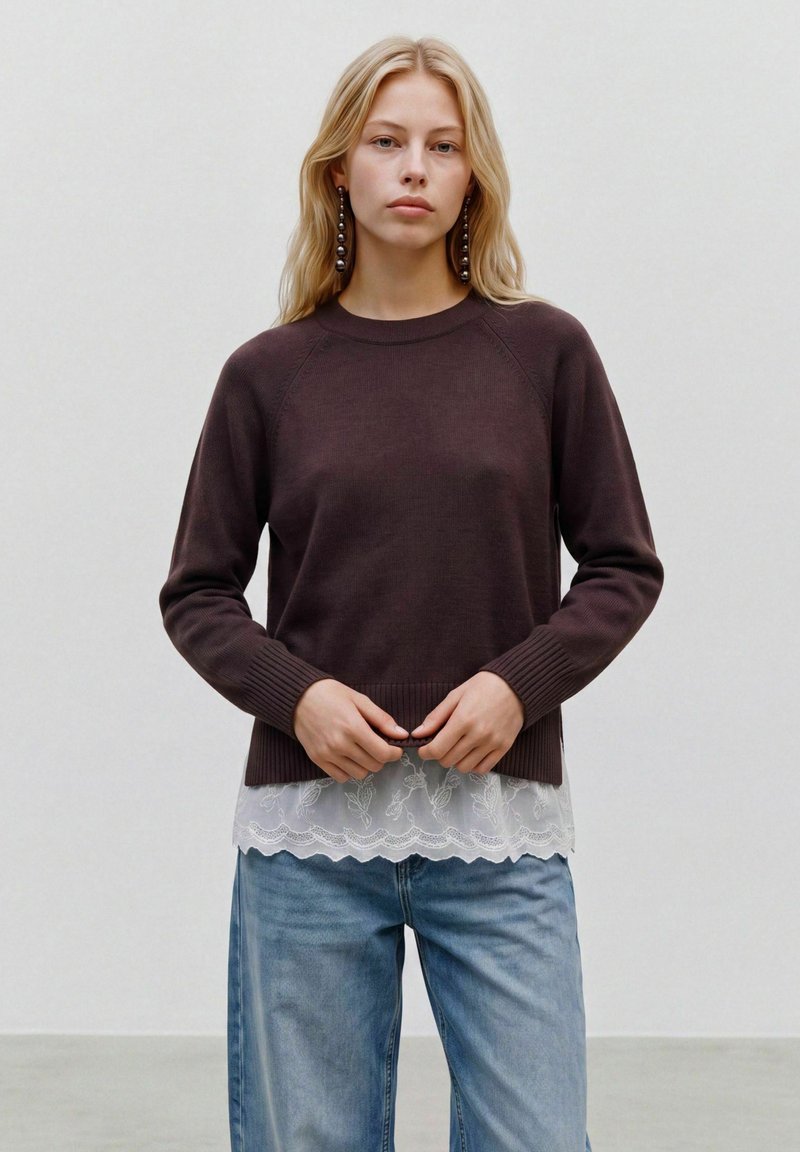 Jeune femme aux longs cheveux blonds porte un pull marron foncé par-dessus un top blanc en dentelle et un jean bleu, debout devant un fond clair uni.