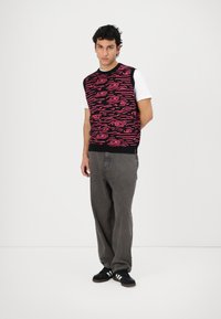 Gebreide mouwloze trui in het zwart met een rood golfpatroon, gedragen over een witte blouse, gecombineerd met grijze broek en zwarte sneakers.