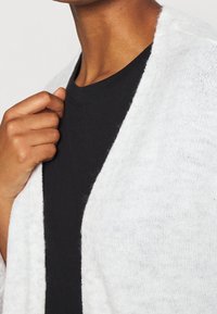 Cardigan bianco, morbido e lavorato a maglia con una vestibilità ampia, indossato sopra una maglietta nera a collo tondo. La texture appare morbida e leggera, con bordi aperti.