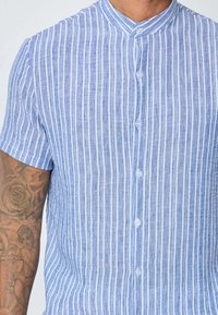 Chemise à manches courtes rayée bleu clair en lin. Rayures verticales de différentes largeurs et design de col à bouton. Dispose de boutons blancs.