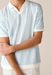 Vaaleansininen kuvioitu poolopaita, jossa on v-pääntie, valkoinen kaulus ja resorireunukset hihoissa. Paita on valmistettu teksturoidusta kankaasta, ja helmassa on yhtenäinen valkoinen reunus.