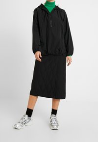 Coupe-vent noir à capuche avec zip à l'avant, associé à une jupe midi matelassée noire. Le modèle porte des baskets de sport argentées et des chaussettes noires.