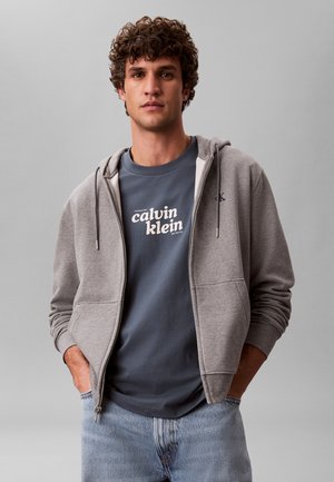 Giovane uomo con capelli ricci che indossa una felpa grigia con zip sopra una t-shirt blu navy Calvin Klein e jeans azzurri, in piedi con espressione neutra su sfondo grigio.