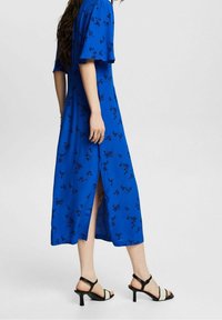 Robe maxi fleurie bleue avec de courtes manches volantées, des fentes sur les côtés et des accents noirs ; portée avec des sandales à talons noires et blanches.