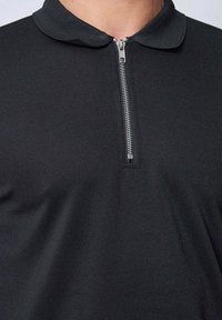 T-shirt polo noir avec un col zippé. Le tissu a un tissage texturé. La fermeture éclair est de couleur argentée et se trouve au centre du cou.