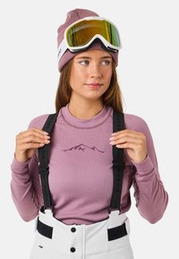 Langsleeve, geribbelde, lichtpaarse basislaag met een bergafbeelding, gecombineerd met witte snowpants en zwarte bretels, afgemaakt met een gebreide beanie en een skibril.