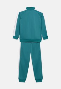 Τυρκουάζ φόρμα με ζακέτα υψηλού γιακά και αντίστοιχο παντελόνι, και τα δύο με λευκές ρίγες στα μανίκια και στα πόδια του παντελονιού.