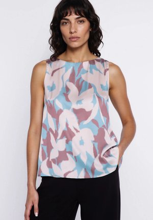 Kobieta o kręconych ciemnych włosach nosi bezrękawnikową, wielokolorową kwiecistą bluzkę w odcieniach niebieskiego, różowego i mauve oraz czarne spodnie, na białym tle.