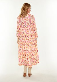 Rochie maxi dintr-un material ușor, cu un model geometric portocaliu și roz, mâneci lungi și un design în straturi. Pur tată cu botine de culoare deschisă.