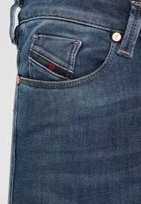 Jeans de mezclilla de un azul oscuro con un pequeño bolsillo frontal, costuras visibles y remaches metálicos cerca de la cintura. Textura suave con un ligero desgaste.