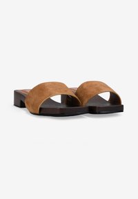 Sandalias marrones de ante con puntera abierta, estilo slip-on, con tacones bajos de bloque y plantillas texturizadas oscuras sobre fondo blanco.