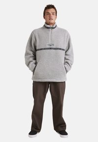 Fleece pullover i lysegrå med lynlås krave, der har et dekorativt bånd i sort og lilla hen over brystet og forlommer.