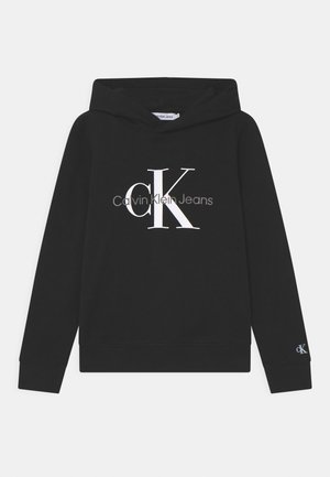 Zwarte capuchontrui met grote witte "cK" en grijze "Calvin Klein Jeans" logo's op de voorkant, en een klein "cK" logo op de linkermouwmanchet.