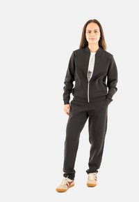 Giacca bomber nera con zip e maniche lunghe, abbinata a pantaloni jogger neri coordinati. Le scarpe sono beige con dettagli rossi e suole testurizzate.